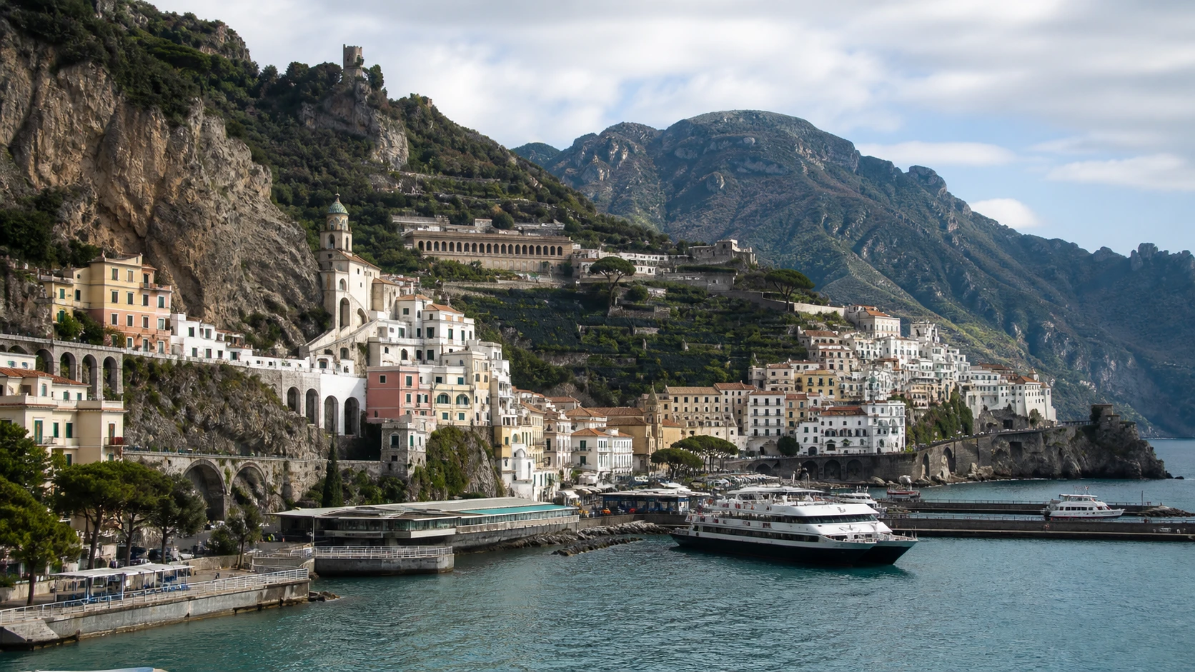 Amalfi town harbor with ferries, waterfront buildings, and steep mountains behind the coast