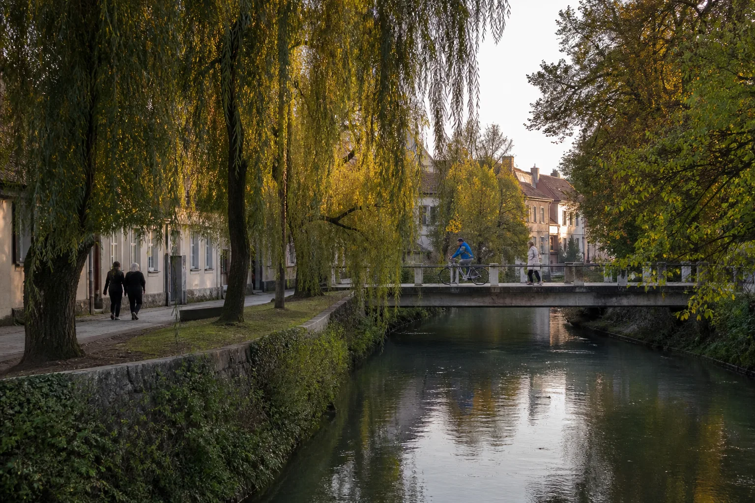 Willow trees, pedestrians, and a cyclist beside the Gradaščica River in Ljubljana's Trnovo neighborhood